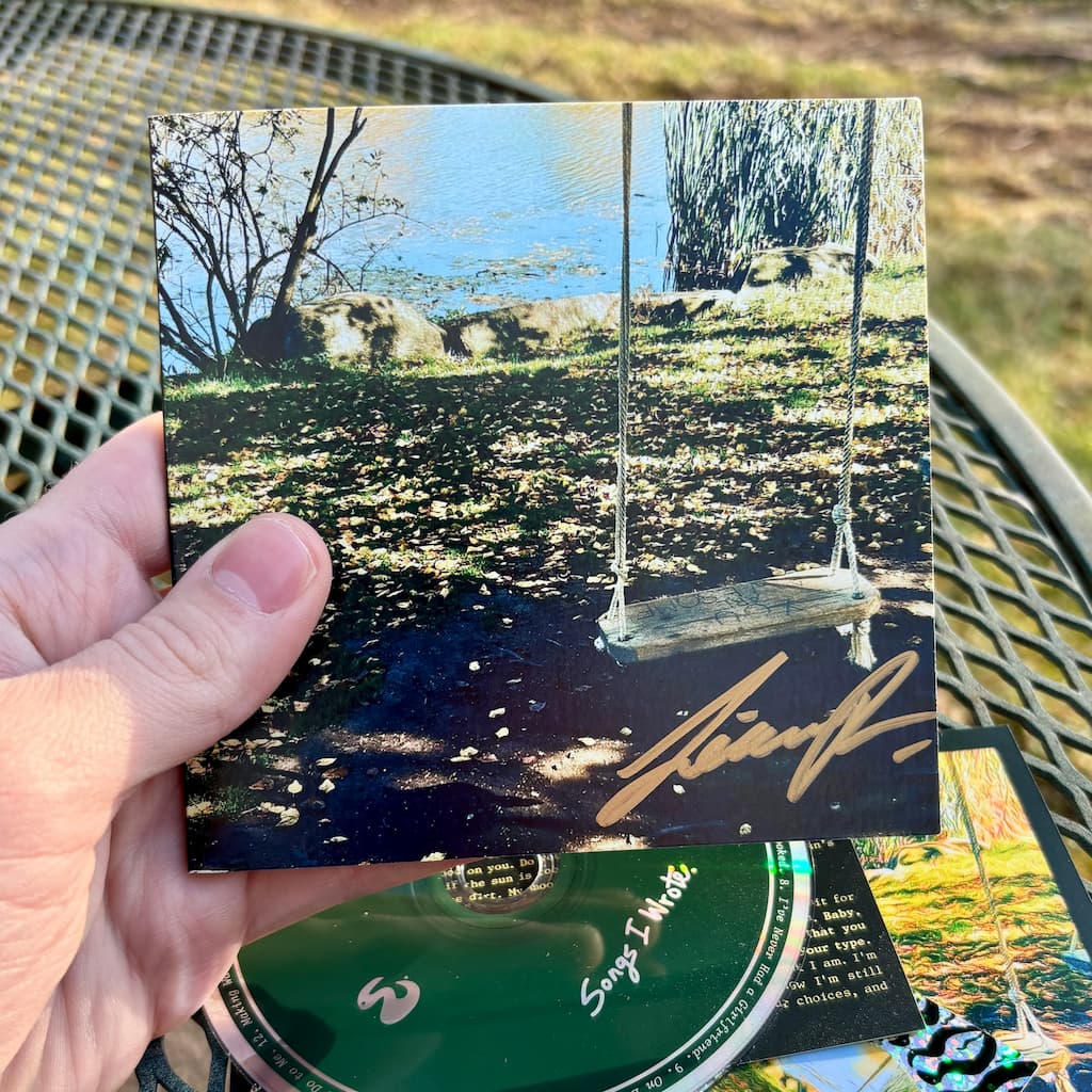 Hand holding a CD wallet with a signature, against a blurred natural background.