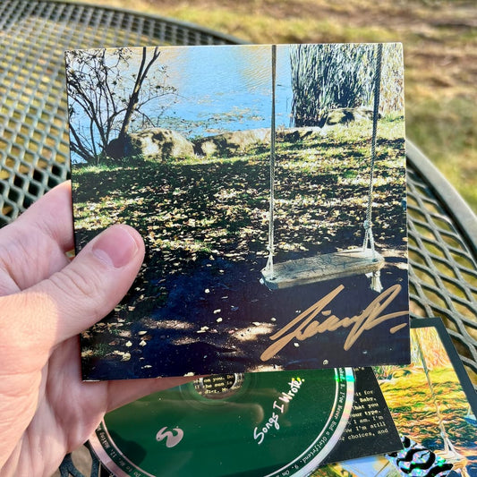 Hand holding a CD wallet with a signature, against a blurred natural background.