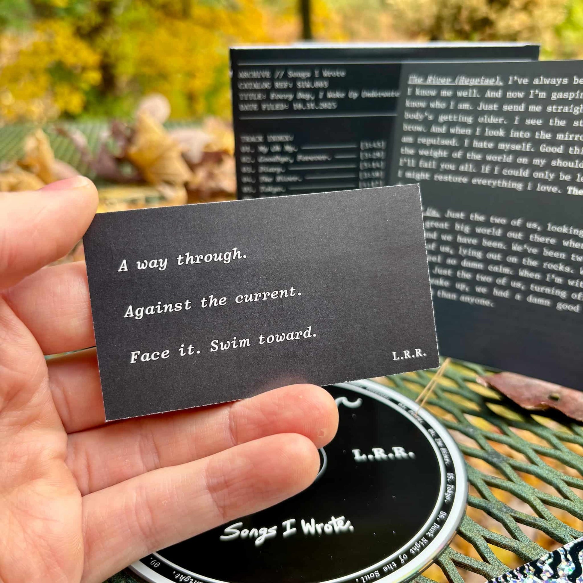 Hand holding a black card with text, with a music album and autumn leaves in the background.