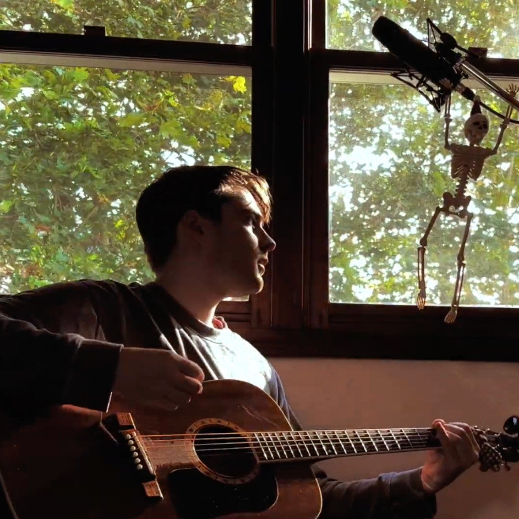 Person playing guitar in a room with a window and skeleton decoration.